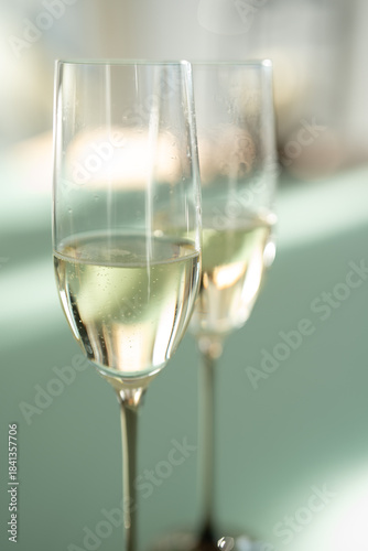 Two filled champagne glasses on a modern glass table. Elegant modern style with soft bright green background und light bokeh. Vertical close-up.