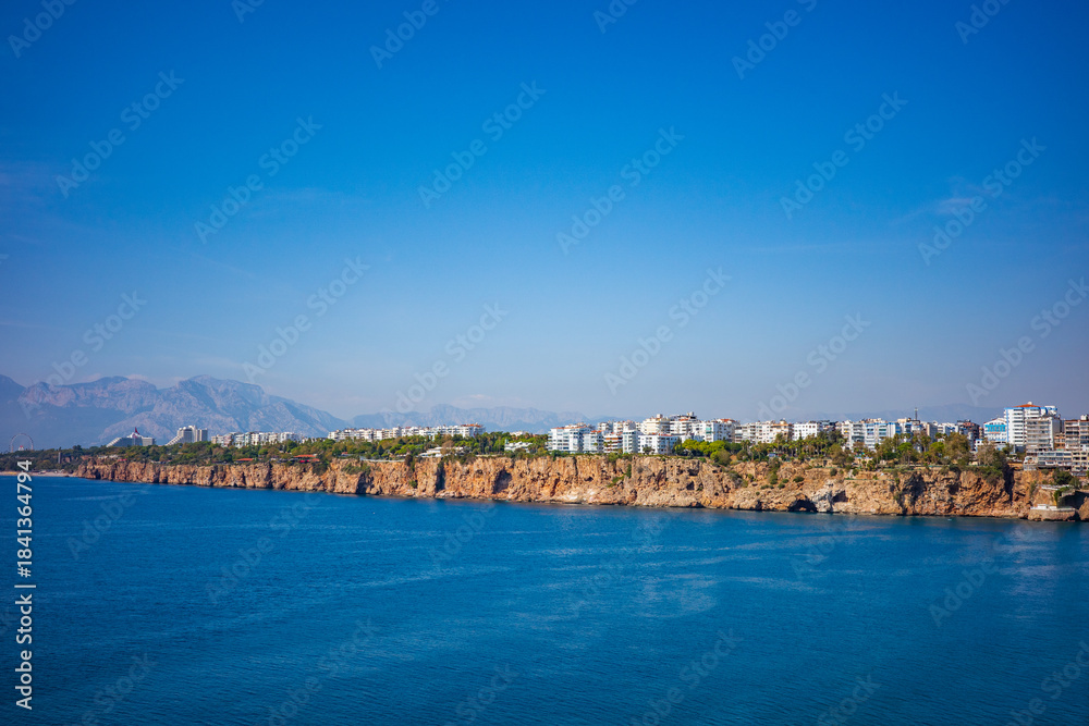 Obraz premium A vibrant panoramic view of Antalya’s rocky coastal cliffs meeting the deep blue Mediterranean Sea, with modern white apartment buildings lining the shoreline and mountains