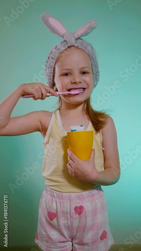 Child with rosy cheeks and a yellow tank top vigorously brushing, showcasing morning routine fun. Close-up portrait of a smiling child using a pink and white toothbrush, fuzzy bunny ears poised above 