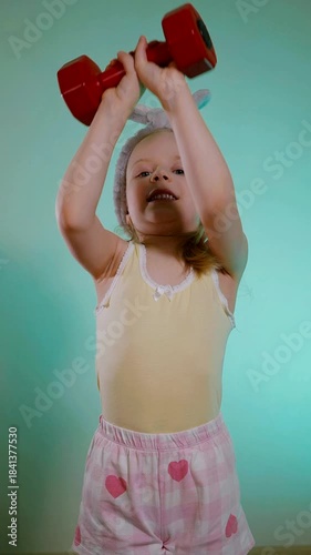 Vertical. Child with wide smile lifting a bright red weight, ready for a fun workout session. Excited young girl wearing bunny ears headband and holding a small red dumbbell against a teal background.