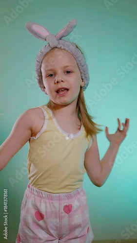Vertical. Young child with bunny ears headband singing and dancing funny against a bright cyan background. Toddler in yellow tank top and pink heart-patterned shorts