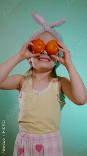 Vertical. Cheerful young girl wearing a fuzzy gray bunny ear headband holds two bright orange tangerines over her eyes, grinning widely against a vibrant teal background.