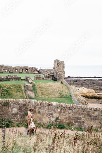 Woman walks near historic stone castle ruins by the sea under cloudy skies in a peaceful coastal scene