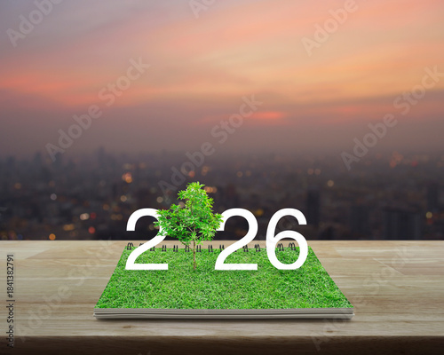 2026 white text with growing tree on green grass on open book on wooden table over blur of cityscape on warm light sundown, Happy new year 2026 ecological cover concept