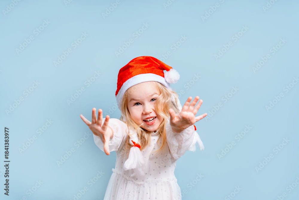 Obraz premium Cheerful little child girl in a white dress and Santa hat on a blue background, the concept of new year and Christmas, space for text on the banner