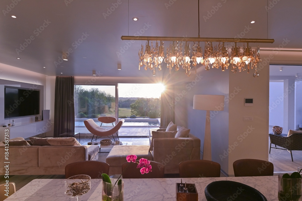 Obraz premium Modern luxury living room interior at sunset. Open-plan space featuring beige sofas, marble dining table, large glass windows with sun flare, and view of a patio. Stylish contemporary home design
