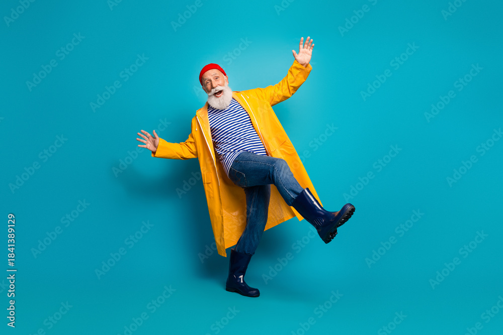 Obraz premium Joyful bearded man in yellow raincoat kicks playfully against blue background