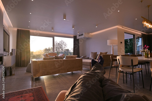 Modern luxury living room interior at sunset. Open-plan space featuring beige sofas, marble dining table, large glass windows with sun flare, and view of a patio. Stylish contemporary home design