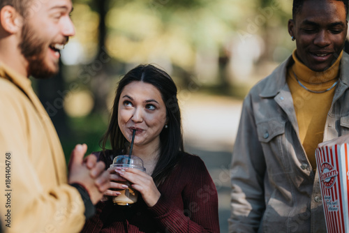 Slika na platnu Three diverse friends chat and laugh in a leafy park, sipping iced coffee while one holds popcorn
