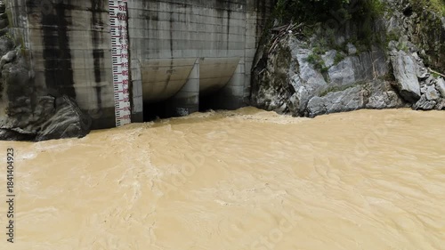 Aerial drone view of a concrete hydropower intake structure on the muddy banks of a flowing river in the lush green mountains of Nepal for renewable energy production project