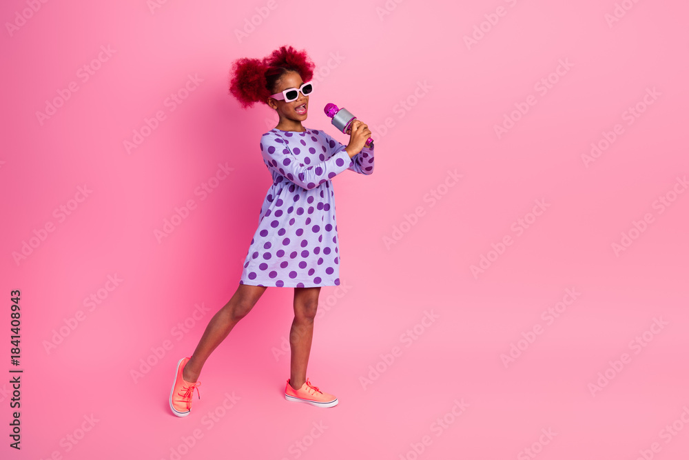 Fototapeta premium Young girl sings into a microphone in a playful pink studio wearing a purple polka dot dress and sneakers