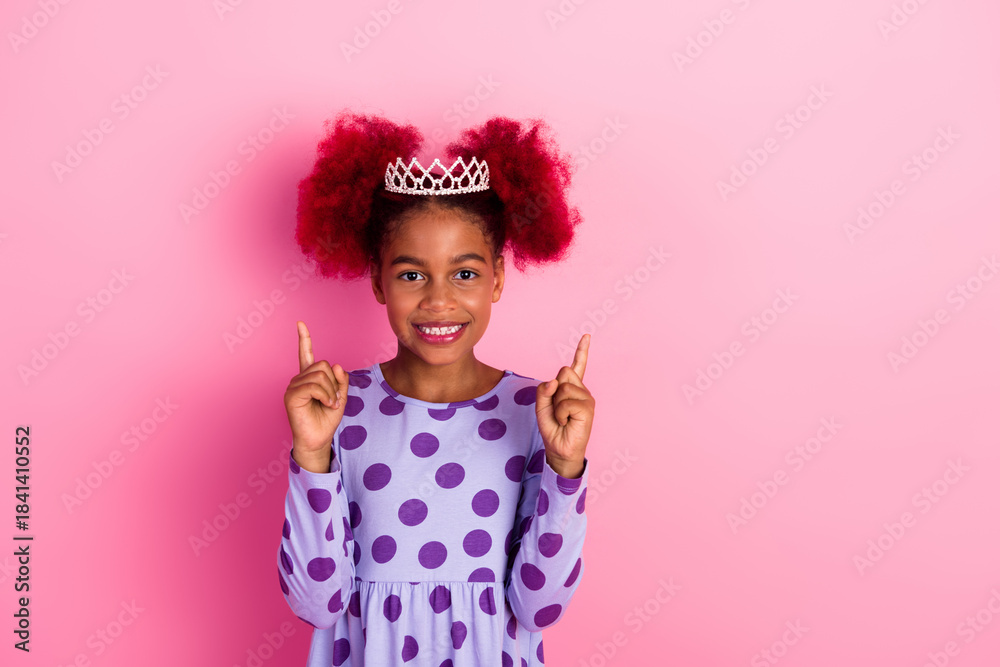 Fototapeta premium Little girl in a purple polka dot dress wears a crown on a pink background smiles and points up