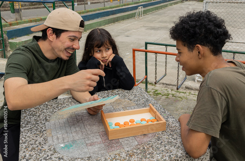 Man with his niece and a friend sitting at a park table playing a board game, enjoying time together outdoors in a cheerful and fun family scene