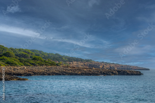 Rugged rocky Mediterranean coastline and dark blue sea with green forest under a cloudy blue sky at sunset with copy space