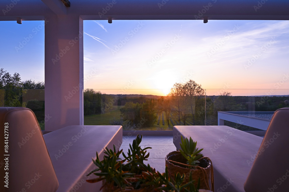 Fototapeta premium Sunset view from a luxurious modern patio with lounge chairs, overlooking a tranquil green landscape of trees and vineyards, perfect for a relaxing evening