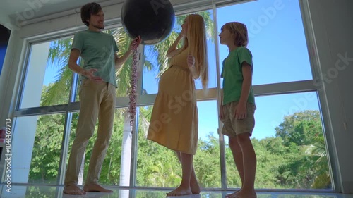 Happy family celebrating indoor gender reveal party at home. Pregnant mother pops balloon with blue confetti announcing it's a boy, while father and elder brother hug, cheer and enjoy joyful family
