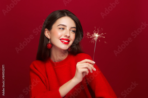 Portrait of beautiful young woman holds a sparkler on red background, red lipstick and festive makeup, Christmas toy earrings, shiny glowing skin, Winter holidays concept