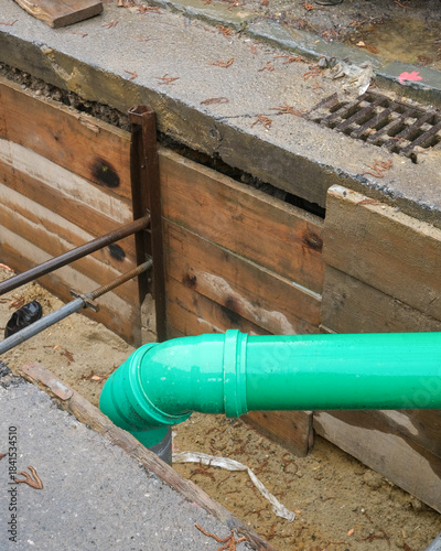 Construction work shows a bright green drainage pipe being installed on a city street surrounded by wooden planks