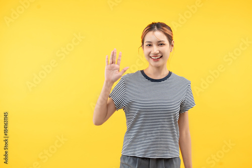 Friendly young asian woman saying hi and waving with a cheerful smile on yellow background.