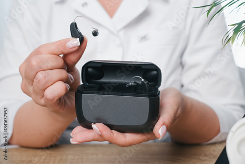 Doctors hands holding an open charging case with two digital hearing aids. Modern assistive technology for hearing health and daily use over white medical uniform