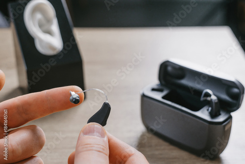Fingers holding tiny digital hearing aid with charging case and ear model on table. Close-up view of assistive hearing technology in audiology clinic