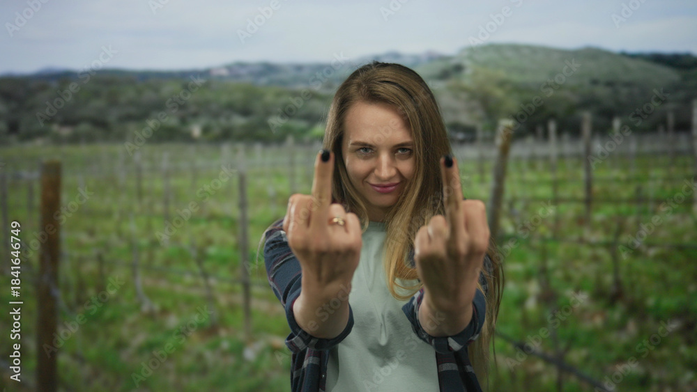 Obraz premium Young woman outdoors in a green field displays a cheeky gesture with both hands, expressing emotion and confidence, wearing casual clothing and smiling playfully on a sunny day.