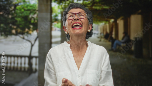 Senior woman with grey hair laughing joyfully in an old town street, creating an engaging scene with her vibrant expression outdoors in an urban setting.