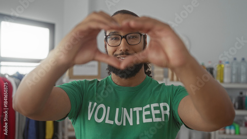 Obraz na płótnie Man in green volunteer shirt wearing glasses forms heart gesture with hands in building donation room with shelves and bottles; compassion community support