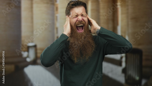 Fotografia Man with long beard screaming with hands raised by historic stone building entrance; anger release solitude