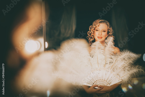 Beauty blonde hair retro lady looking into mirror hand holding fan of natural white ostrich feathers. sexy woman burlesque show actress in dressing room. Flapper girl 20s roaring twenties old style