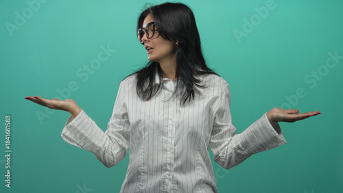 фотографія Young hispanic woman wearing glasses and striped shirt holding open palms in a teal studio; uncertainty