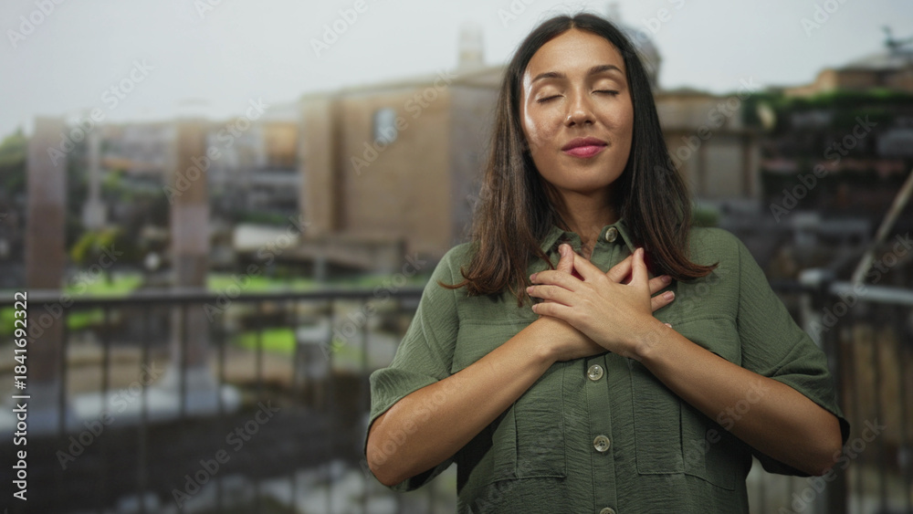 Naklejka premium Woman with hands crossed over chest on building balcony, closed eyes and gentle smile; gratitude reflection.