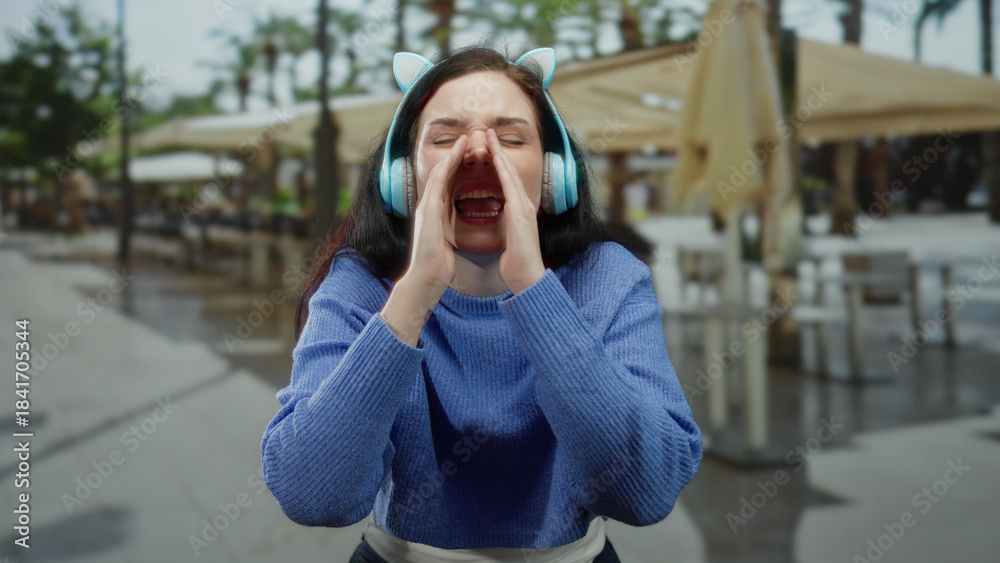 Fototapeta premium Woman wearing headphones shouting playfully on a terrace with blurred street and restaurant in the background showcasing a lively and casual outdoor scene.