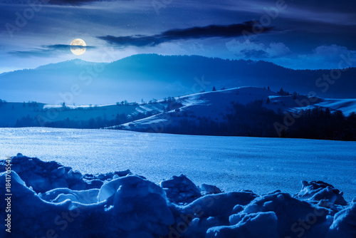 beautiful winter landscape with snow covered rolling hills under dark sky at night. majestic mountains in full moon light. frosty countryside scenery. backdrop for fiction art or mystery concepts