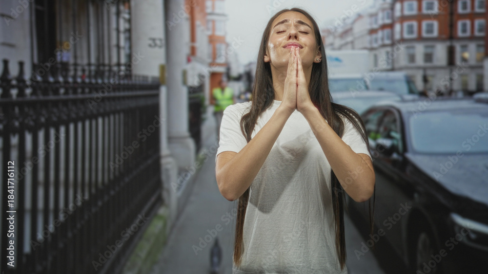 Fototapeta premium Woman with hands clasped praying on a street in a city near parked cars and an iron fence; reflection hope serene.