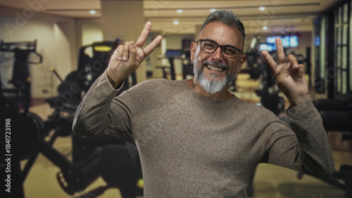 Billede på lærred Man middle aged with beard and glasses flashing peace sign with both hands while smiling in gym building; joy wellness