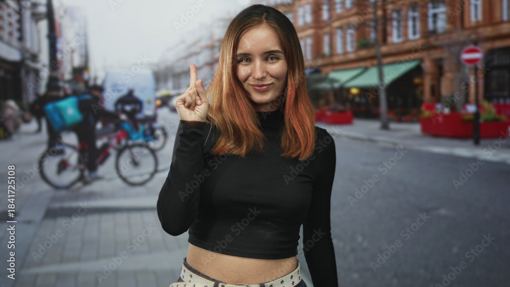 Obraz premium Redhead woman smiling points finger upward on a busy urban street beside a cyclist and a parked car, facing camera in a cropped midshot; confidence.