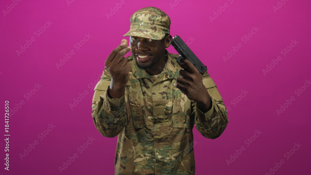 Fototapeta premium Man soldier in camouflage uniform holding a gun to his head while making a pinching fingers gesture against a magenta studio backdrop, smiling; tension threat.