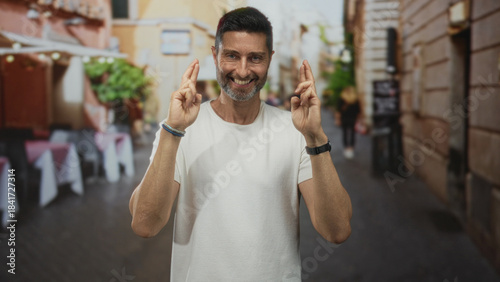 Bilde på lerret Man shows crossed fingers with both hands while smiling on street outside restaurant terrace; hopeful anticipation
