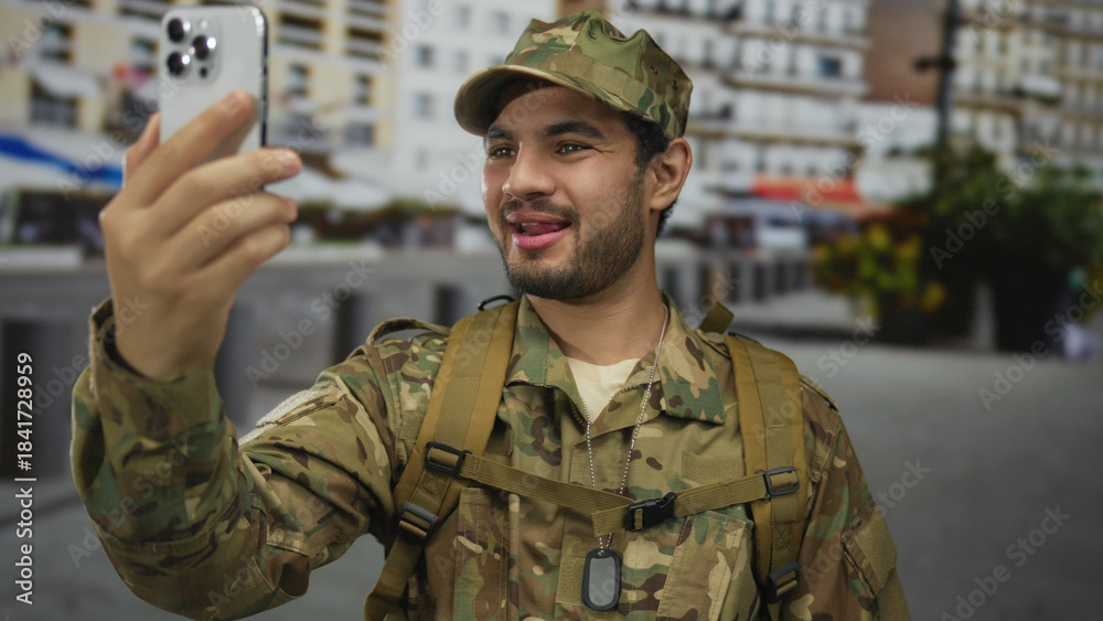 Fototapeta premium Man in military uniform holds smartphone for a video call on a busy urban street lined with apartment buildings; connection communication camaraderie service.