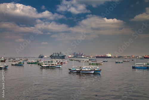 View of a fleet of small fishing boats bobbing gently on the water, with a distant cargo ship and cranes under a partly cloudy sky, Karachi, Sindh, Pakistan.