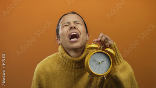 Hispanic woman in a yellow sweater holds a yellow alarm clock against an orange background, expressing frustration over time management issues.