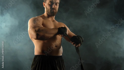 A muscular fighter wraps bandages around his hands on a black background in the smoke