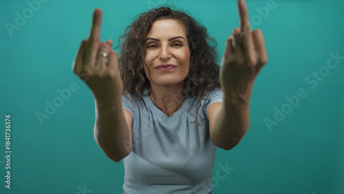 Obraz na plátně Hispanic woman with curly hair and blue t shirt extends middle fingers in teal studio environment; defiance