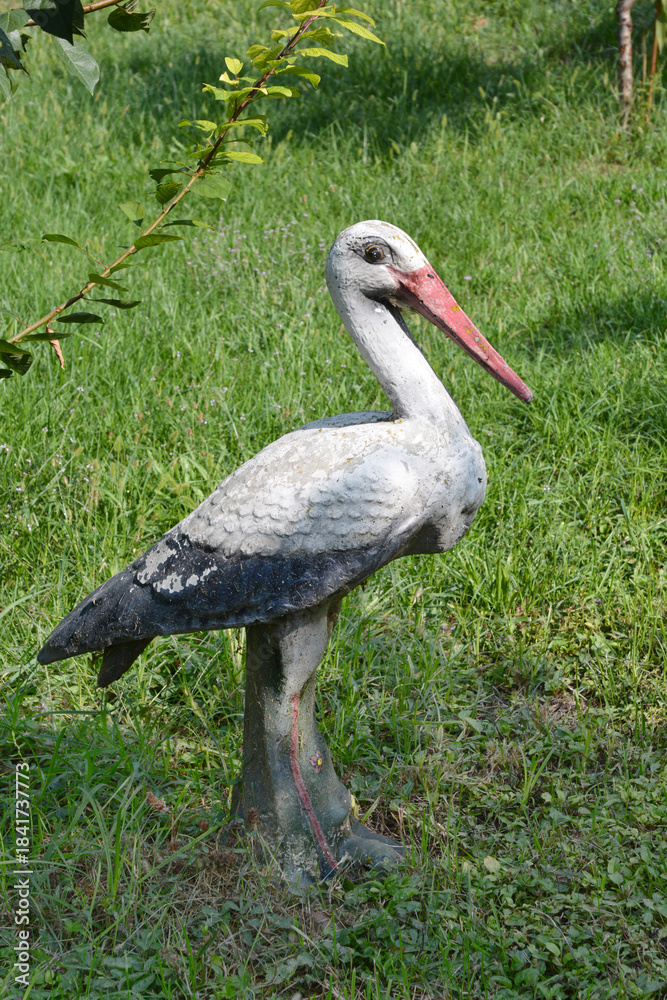 Obraz premium A weathered, white and black garden sculpture of a stork stands on a patch of green grass in bright sunlight