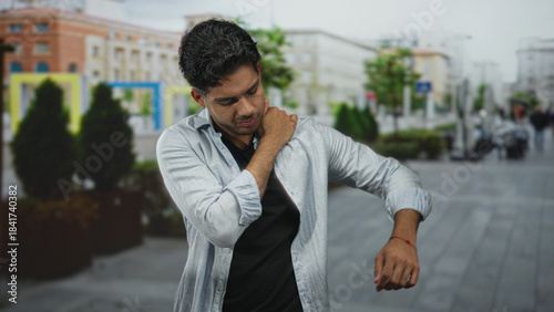 Young hispanic man rubbing neck and clutching shoulder while standing on a busy city street; discomfort fatigue soreness recovery.