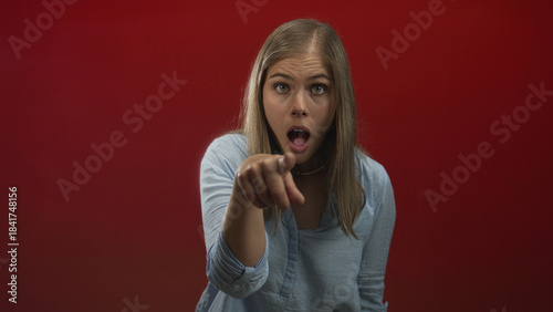 Young blonde woman points finger at camera in studio with red wall, mouth open and direct gaze; surprise accusation.