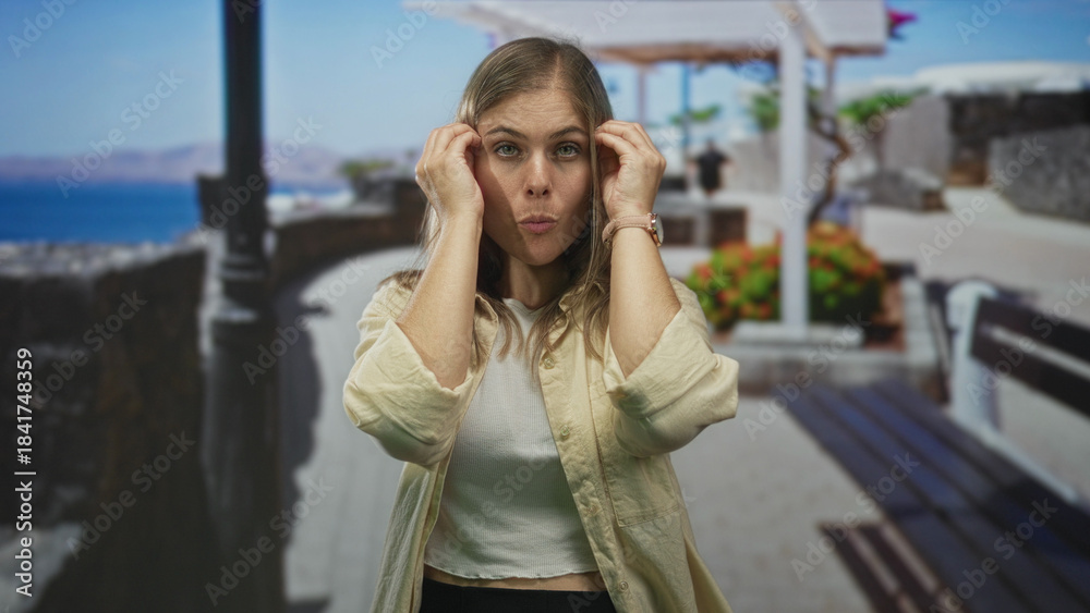 Fototapeta premium Woman with hands pressing temples in bright seaside street wearing casual attire and thoughtful posture; calm reflection introspection mood.