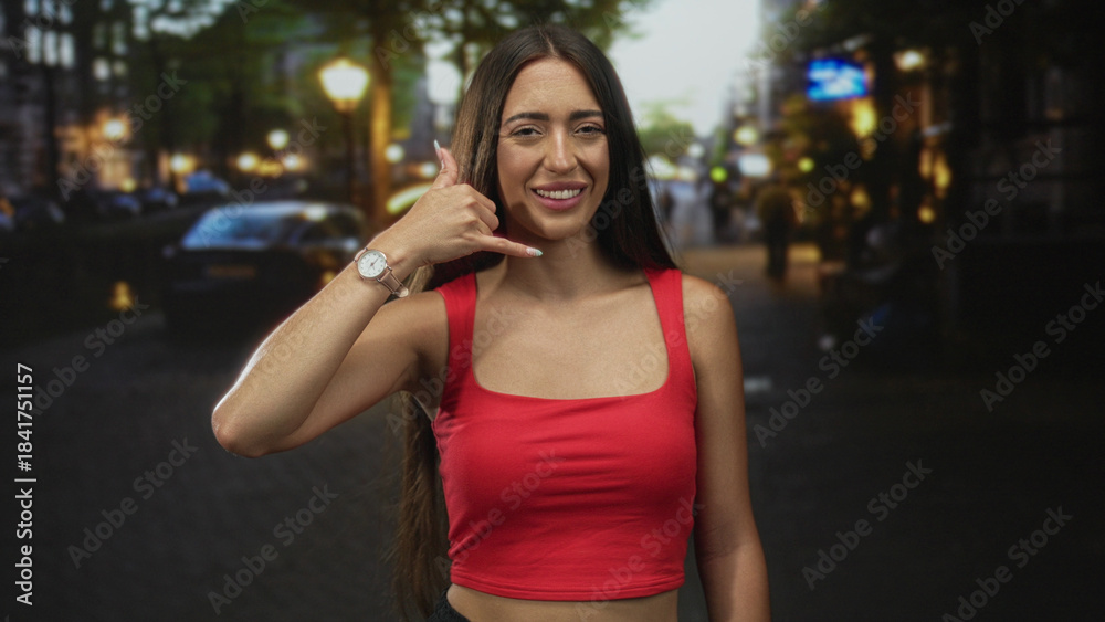 Fototapeta premium Woman smiling and making call gesture on city street in red crop top with visible watch and bare shoulders; playful social invitation.