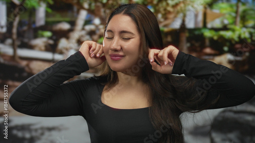 Young hispanic woman plugs ears with fingers in lush green park surrounded by blurred trees and sunlight overhead; annoyance.